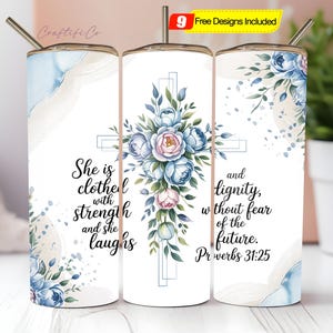 May include: A white tumbler with a floral cross design, featuring blue and pink flowers and green leaves. The tumbler has the text "She is clothed with strength and she laughs" and "and dignity, without fear of the future. Proverbs 31:25". Includes a straw.