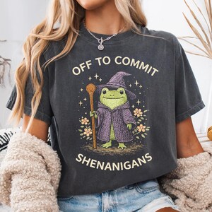 Op de afbeelding: Donkergrijs t-shirt met de tekst "OFF TO COMMIT SHENANIGANS". De afbeelding toont een groene kikker met een paarse tovenaars hoed en mantel, die een houten staf vasthoudt, omringd door bloemen en sterren.