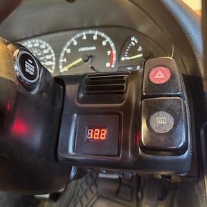May include: A black car dashboard console featuring a digital red LED voltage display, a round engine start stop button, a hazard light switch, and a rear window defroster button, positioned in front of an analogue speedometer and tachometer gauge cluster.