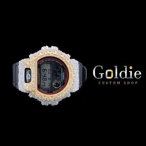 Könnte beinhalten: Eine schwarze Digitaluhr mit einer gold- und diamantbesetzten Lünette. Das Zifferblatt zeigt die Uhrzeit und das Datum an. Der Markenname "Goldie Custom Shop" ist in Silber dargestellt.
