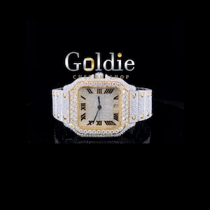 May include: A luxury wristwatch with a square face, featuring a gold-tone bezel and a silver-tone band. The watch is encrusted with numerous small, sparkling stones. The watch face displays Roman numerals and the brand name "Goldie Custom Shop" is visible.