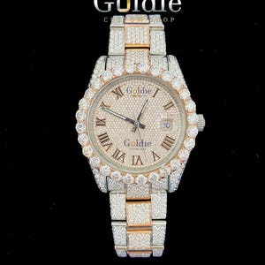 May include: A luxury wristwatch with a silver and gold strap and a diamond-encrusted bezel. The watch face is a light champagne colour with Roman numerals and the brand name "Goldie". The watch has a date display at the 3 o'clock position.