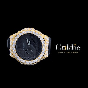 Puede incluir: Un reloj G-Shock negro con un bisel dorado incrustado con piedras preciosas transparentes. La esfera del reloj es negra con manecillas y marcadores blancos. El reloj está sobre un fondo negro, con el texto "Goldie Custom Shop" en la esquina inferior derecha.
