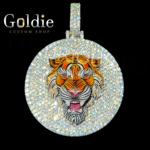 May include: A circular, diamond-set pendant showcasing a tiger's head with orange, white, and black stripes. The tiger's face is central, with an open mouth and pink tongue. The pendant has a silver bail and the text "Goldie Custom Shop".