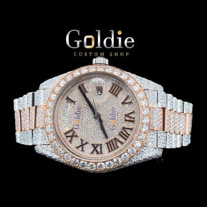 May include: A diamond-encrusted wristwatch with a silver and rose gold strap. The watch face has Roman numerals and the word "Goldie". The bezel and face are covered in diamonds. The background is black.