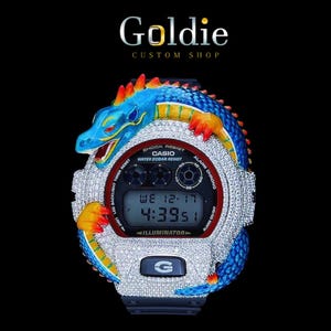Könnte beinhalten: Eine individuelle Casio G-Shock Uhr mit einem Drachendesign. Die Uhr hat einen blauen, orangefarbenen und gelben Drachen, der sich um das mit Kristallen besetzte Zifferblatt windet. Das Zifferblatt zeigt Datum und Uhrzeit an. Der Text "Goldie Custom Shop" steht oben.