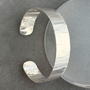 May include: A polished silver cuff bracelet featuring a wide band with a series of subtle, vertical indentations along the outer surface. The bracelet has an open design with rounded ends, showcasing a reflective, metallic finish throughout.