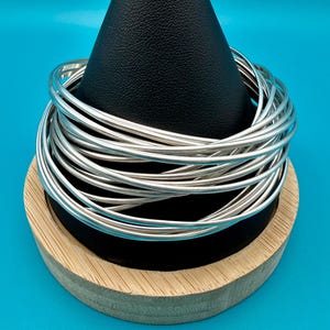 May include: A stack of silver-colored bangles displayed on a black cone-shaped stand atop a wooden base. The bangles are thin and round, and the stand is set against a bright blue background. The jewelry is simple and elegant.