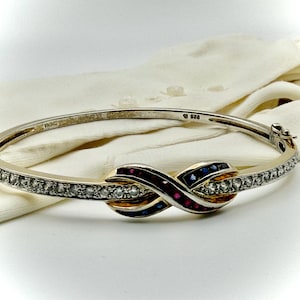 May include: A silver-tone hinged bangle bracelet featuring an infinity-shaped center design. The band is inlaid with clear stones, while the center infinity loop displays alternating rows of red and blue stones. The interior is stamped 925.