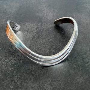 May include: A silver-toned metal cuff bracelet featuring a triple-band design with a smooth, polished finish. The bracelet has an open-ended construction and a curved, wave-like shape that creates a layered appearance across the wrist area.