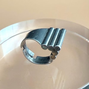 May include: A silver-toned metal ring featuring an open band design. The top of the ring is composed of three parallel, cylindrical bars arranged in a stepped formation, creating a geometric structure with a smooth, metallic finish throughout.