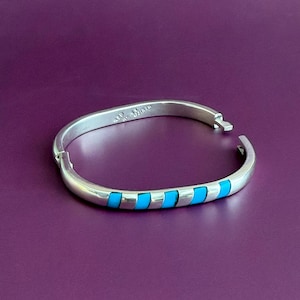 May include: A silver hinged bangle bracelet featuring a series of inlaid rectangular turquoise-colored segments along the front section. The interior of the metal band displays stamped text including the word Mexico and other markings.