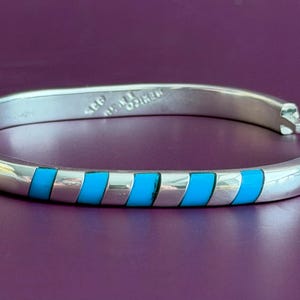 May include: A silver hinged bangle bracelet featuring a series of inlaid bright turquoise rectangular segments arranged in a diagonal pattern. The interior of the metal band is stamped with the text 925 and MEXICO, indicating its composition.