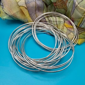 May include: A collection of silver-colored bangles, arranged in a circular pattern, are displayed on a bright blue surface. A colorful scarf with green, yellow, and purple hues is in the background. The bangles are thin and shiny.
