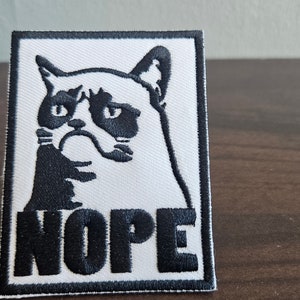 Op de afbeelding: Een witte geborduurde patch met een zwart-witte afbeelding van een chagrijnige kat en het woord "NOPE" in zwart.
