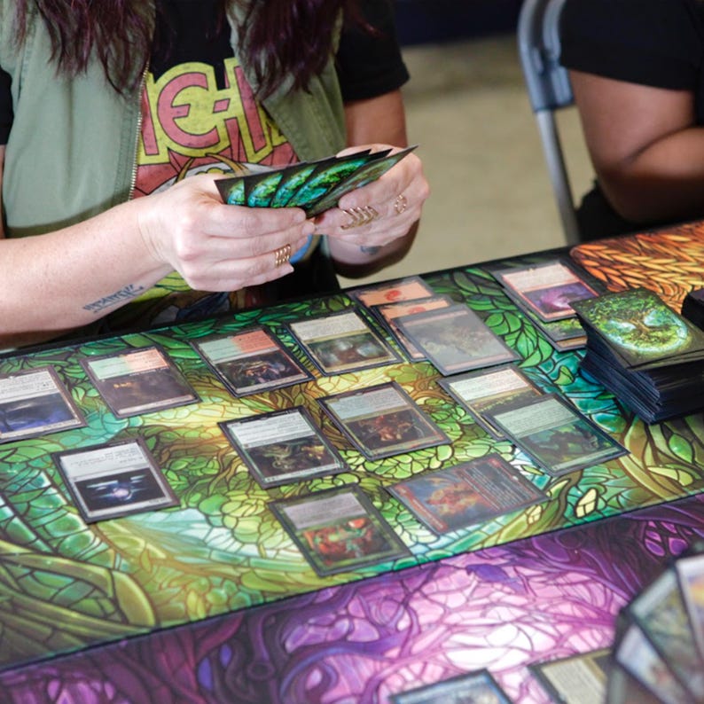 May include: A person is playing a card game with a green and purple game mat. The mat has a stained glass design. The person is holding a stack of cards in their hands. There are many cards laid out on the mat.