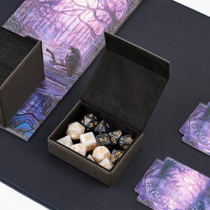 May include: A black felt dice box with a hinged lid, open to reveal a collection of white and black dice with gold numbers. The dice are arranged in a random pattern inside the box.