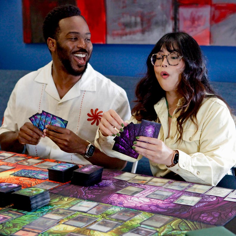 May include: Two people are playing a card game with a large, colorful game board. The game board has a purple, green, and blue design. The people are holding cards with a purple, green, and blue design.