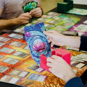 May include: A colorful deck box with a floral design and a pink lotus flower. The box is made of cardboard and has a hinged lid. It is sitting on a table with a variety of playing cards.