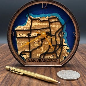 May include: A wooden clock featuring a detailed map of San Francisco, with the city's name at the base. The clock face is blue with a wooden frame, and the hands are gold. A gold pen and a coin are next to the clock.