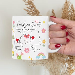 May include: White ceramic mug with the text "I wish we lived closer." The mug features state outlines with hearts, a butterfly, flowers, and a rainbow. Names "Hanna" and "Maria" are below the states. A perfect gift for long-distance friends.