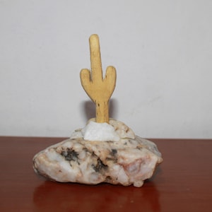Puede incluir: Una escultura decorativa de cactus. Un cactus de madera de color claro está montado sobre una base de piedra moteada, beige y blanca. El cactus es de color marrón claro y está de pie. La base de piedra tiene una pequeña piedra blanca en la base del cactus.