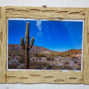 Puede incluir: Una impresión enmarcada de un paisaje desértico con un gran cactus saguaro bajo un cielo azul brillante. El marco es de madera clara con un diseño texturizado único, que añade una estética natural a la obra de arte. La imagen captura la esencia del desierto.