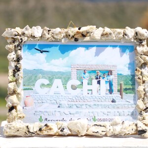 Puede incluir: Un marco de fotos con un borde decorativo de pequeñas piedras claras. La foto presenta la palabra "CACHI" en letras blancas grandes, con personas detrás. El marco descansa sobre una superficie blanca.