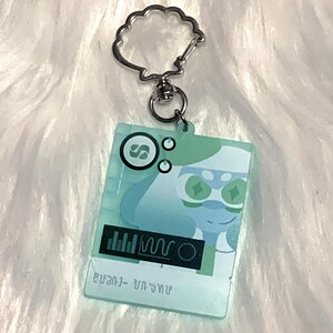 May include: A light blue and green character keychain with a silver key ring. The character has green eyes and a white face. The keychain has a black and white graphic of a sound wave and the text "звук - звук".