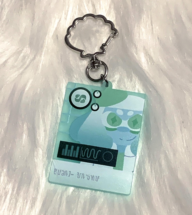 May include: A light blue and green character keychain with a silver key ring. The character has green eyes and a white face. The keychain has a black and white graphic of a sound wave and the text "звук - звук".