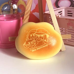 May include: A heart-shaped, yellow-orange squishy toy with a cartoon graphic and the text "NEW SQUIDBEAK". The toy is on a white surface, with a pink gumball machine and a pink basket in the background.