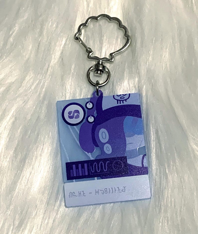 May include: A blue and purple character with a skull on its head is featured on a keychain. The character is holding a microphone and has a music note symbol on its chest. The keychain is made of clear acrylic and has a silver metal ring.