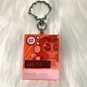 May include: A red and pink acrylic keychain featuring a cartoon character with large eyes and a pink nose. The character is wearing a red and white striped shirt. The keychain has a silver metal ring and a text label that reads "Anya 8 - Psyduck".