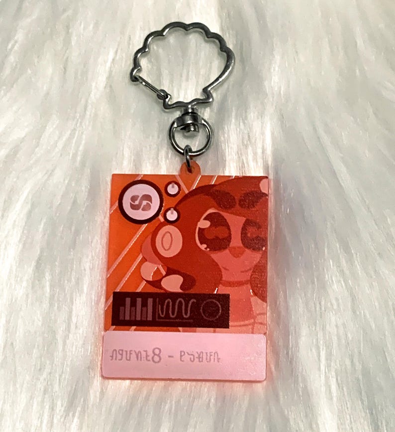 May include: A red and pink acrylic keychain featuring a cartoon character with large eyes and a pink nose. The character is wearing a red and white striped shirt. The keychain has a silver metal ring and a text label that reads "Anya 8 - Psyduck".
