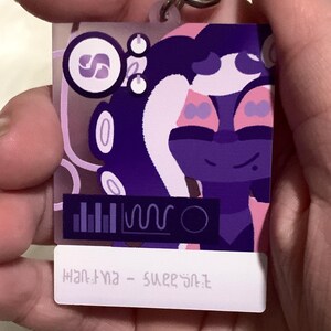 May include: Purple and pink cartoon character keychain with a white background. The character has a purple body, pink hair, and a white face with purple eyes. The keychain also features a small purple and white logo with the letter "S" in the center. The text "HANTYNA - SUPERSTAR" is printed on the bottom of the keychain.