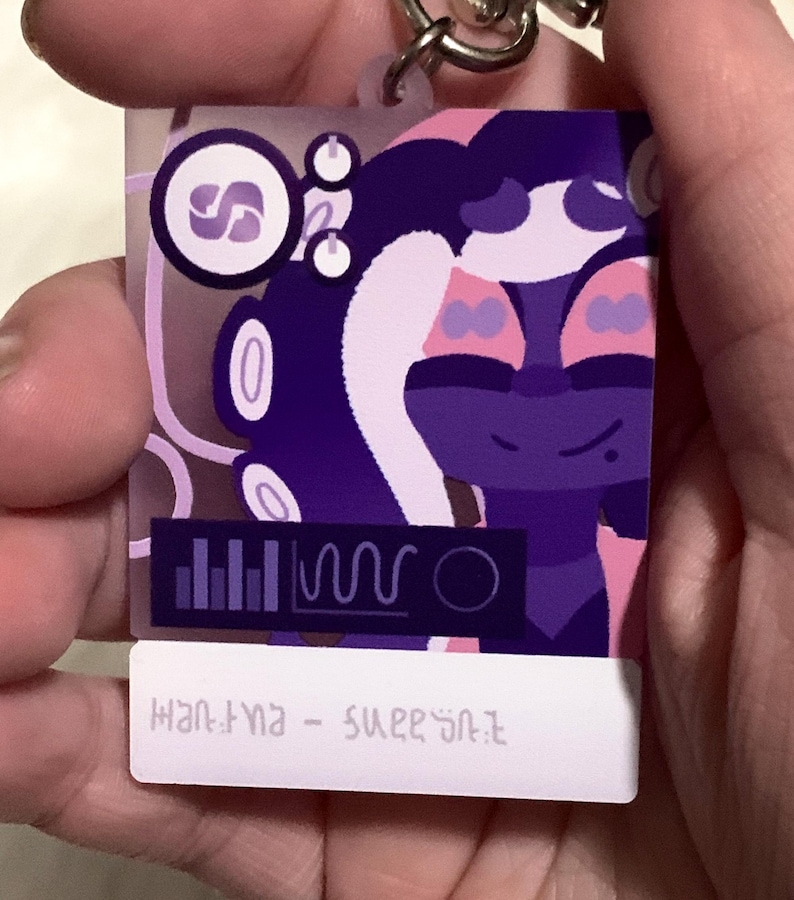 May include: Purple and pink cartoon character keychain with a white background. The character has a purple body, pink hair, and a white face with purple eyes. The keychain also features a small purple and white logo with the letter "S" in the center. The text "HANTYNA - SUPERSTAR" is printed on the bottom of the keychain.