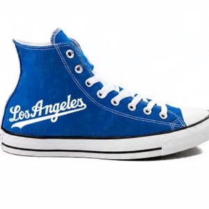 Puede incluir: Una zapatilla de caña alta de lona azul brillante con el texto Los Angeles en blanco en el lateral. El calzado incluye una puntera de goma blanca, cordones blancos, ojales de metal plateado y una suela de goma blanca con una fina raya negra.