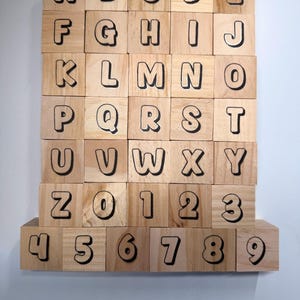 May include: Wooden alphabet and number blocks. Each block is a light brown color with a black outline. The letters A through Z and numbers 0 through 9 are displayed. The blocks are arranged in a grid pattern.