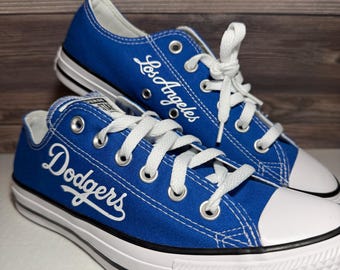 May include: A pair of royal blue low-top canvas sneakers with white laces and white rubber toe caps. The side of one shoe features the white embroidered text "Dodgers" in a script font, while the other displays "Los Angeles" in white script.