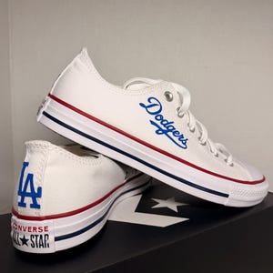 Pode incluir: Tênis Converse branco de cano baixo com a palavra "Dodgers" em azul na lateral. Os tênis têm listras vermelhas e azuis ao longo da sola de borracha branca e o logotipo LA no calcanhar.