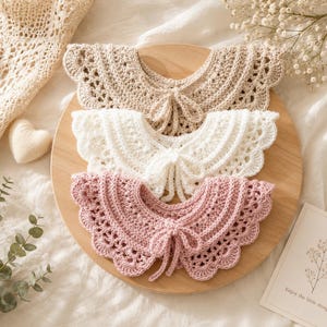 May include: Three crocheted collars in beige, white, and pink, each with a scalloped edge and a bow at the neckline. The collars are arranged on a wooden surface, with a soft, neutral background.