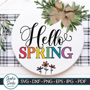 May include: A white round wooden sign with the text "Hello Spring" in black cursive lettering. The word "Spring" is in rainbow colors. There are three small black flowers with green stems below the text.