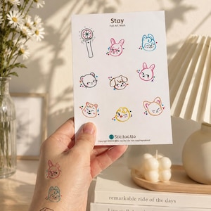 Op de afbeelding: Een wit stickervel met de titel Stay Fan Art Work met negen kleurrijke cartoonkarakterillustraties en een lightstick-pictogram. Drie van de karakterontwerpen zijn ook als tijdelijke tatoeages op een hand aangebracht.