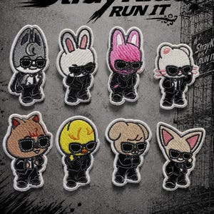 May include: Eight embroidered iron-on patches featuring cartoon characters wearing black suits and sunglasses. The text reads IRON ON PATCH Stray Kids RUN IT. Each character has a distinct animal-like appearance in various colours including grey, white, pink, brown, and yellow.