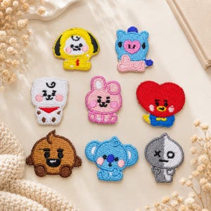 Puede incluir: Un conjunto de ocho parches bordados con personajes de dibujos animados coloridos. El set incluye un perro amarillo, un oso azul, una criatura blanca con bufanda roja, un conejo rosa, un corazón rojo, una criatura marrón, un koala azul y un fantasma gris.