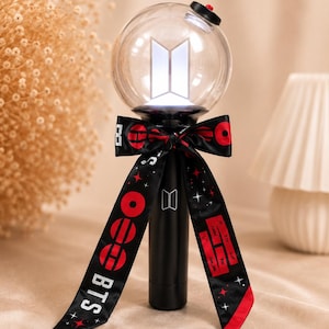 Può includere: Un bastone luminoso BTS nero con una parte superiore sferica trasparente contenente un logo bianco. L'impugnatura è decorata con un nastro di raso nero stampato con motivi circolari rossi, stelle bianche e il testo BTS. Un piccolo tasto rosso è in cima.