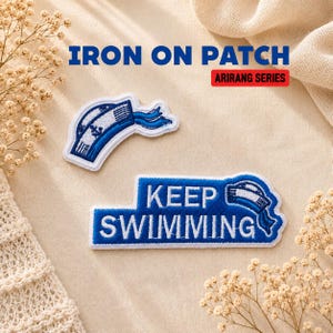 May include: Two embroidered iron-on patches from the Arirang Series. One patch is a blue and white sailor hat with an anchor icon. The second rectangular patch features the blue text KEEP SWIMMING with a small matching sailor hat graphic.