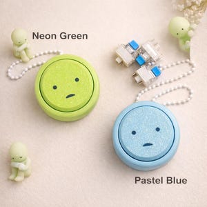 Puede incluir: Llaveros redondos verde neón y azul pastel con un diseño de cara triste. Cada llavero tiene una cadena de cuentas del mismo color y una pequeña figura. El texto "Neon Green" y "Pastel Blue" también es visible.
