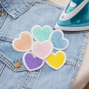 May include: A selection of heart-shaped embroidered patches in pastel shades of peach, mint, pink, blue, purple, and yellow, each with a white border. The patches are placed on a light blue denim jacket, with a teal iron in the background.