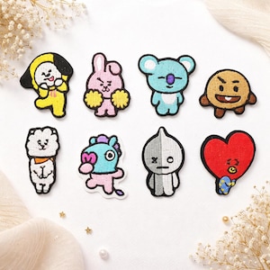 Könnte beinhalten: Eine Sammlung von acht gestickten Aufnähern mit Cartoon-Charakteren in verschiedenen Farben. Die Aufnäher zeigen eine gelbe Figur mit schwarzer Kapuze, ein rosa Kaninchen mit Bommeln, einen blauen Koala, eine braune Figur, einen weißen Löwen, ein blaues Pferd, eine graue Figur und ein rotes Herz.